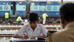 परीक्षा लिखता हुआ छात्र, पृष्ठभूमि में रेलवे से जुड़ी गतिविधियां — परीक्षा और रोजगार से जुड़े समाचार का प्रतीकात्मक दृश्य